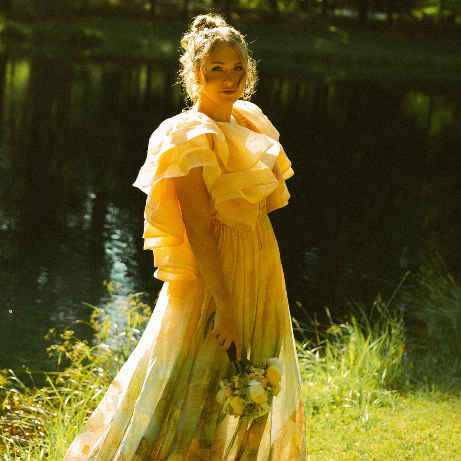 Woman in a yellow dress standing by a pond with trees in the background